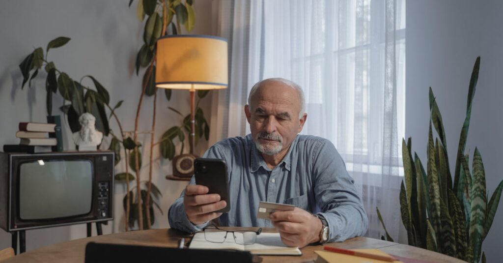 An elderly man sits at a table with a smartphone and credit card, making an online purchase.