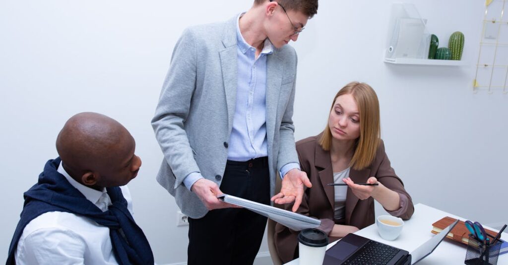 Three business professionals engage in a discussion at an office desk with laptops and coffee.